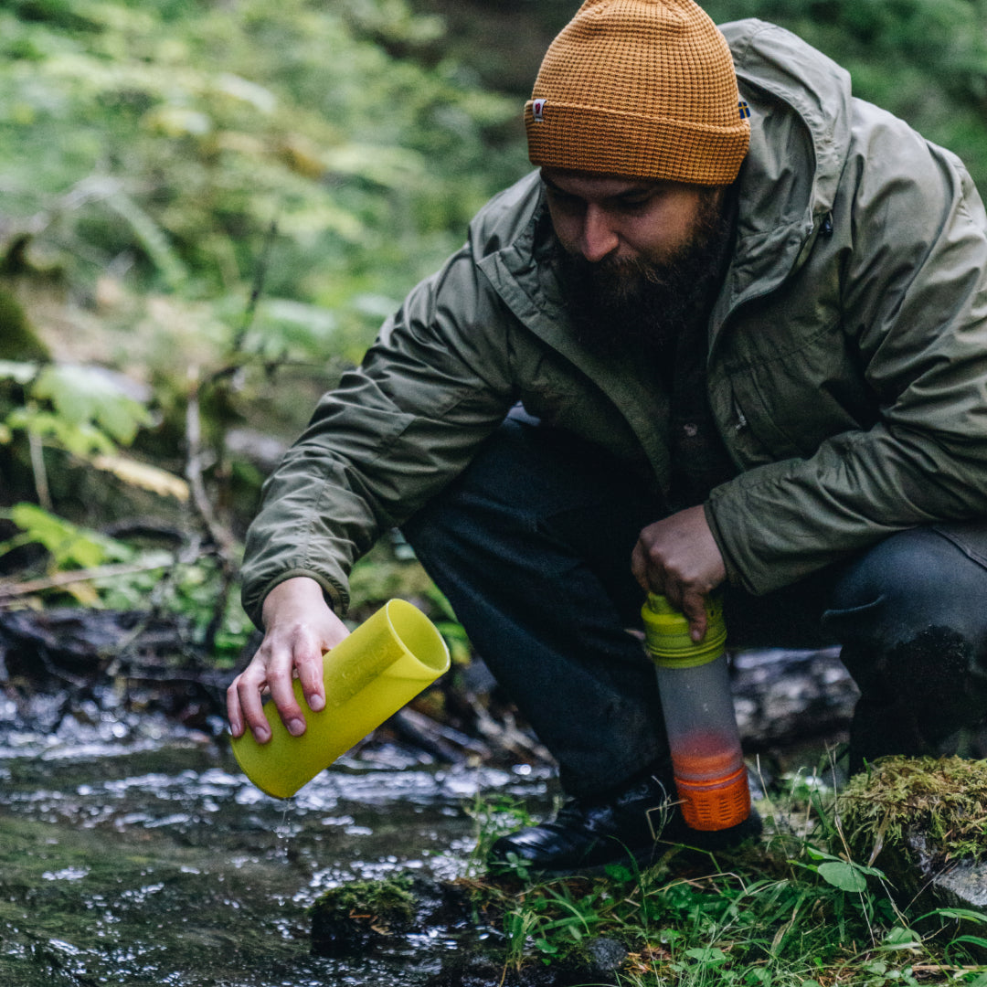 man squatting down to fill his Grayl from a shallow stream